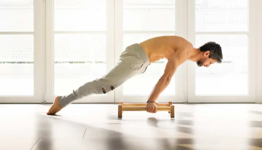 Hombre haciendo flexiones en paralelas de madera en un suelo claro, con luz entrando por ventanas al fondo.