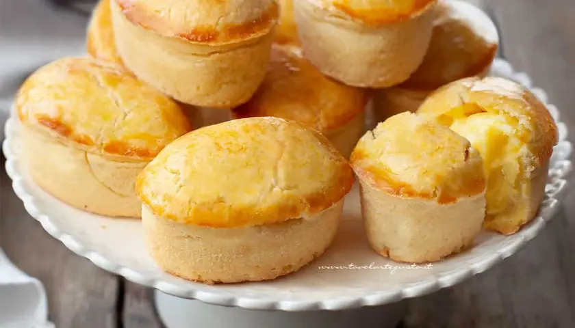 Un mucchio di pasticciotti leccesi dorati e invitanti su un'alzata bianca, pronti per essere gustati.