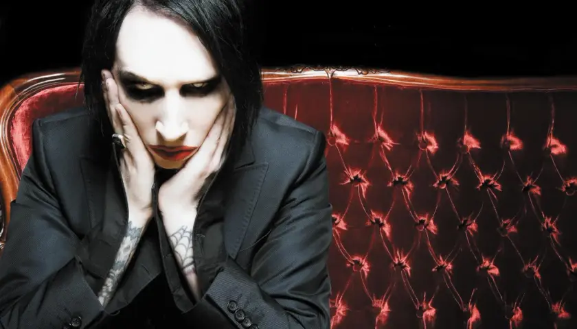 Marilyn Manson, z charakterystycznym makijażem, siedzi na aksamitnej kanapie. Jego utwory często poruszają mroczne tematy.
