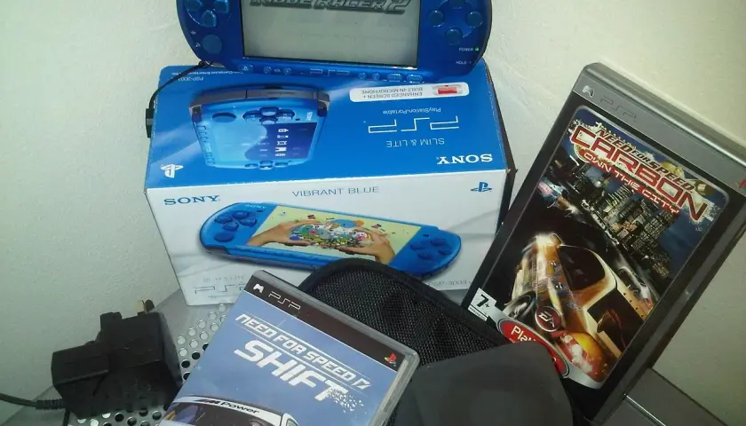 Niebieska konsola PSP z grą Ridge Racer 2, pudełko z konsolą i grami Need for Speed Shift i Carbon. Klasyczne stare gry na telefon.