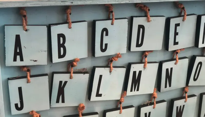 Drewniane tabliczki z literami alfabetu angielskiego pisanego, wiszące na sznurkach. Widoczne litery A, B, C, D, E, J, K, L, M, N, O, U, V, W.