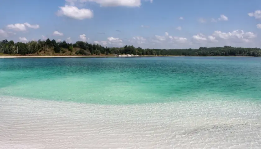 Lazurowe jezioro z piaszczystą plażą i lasem w tle. Woda jest krystalicznie czysta, a dno widoczne.