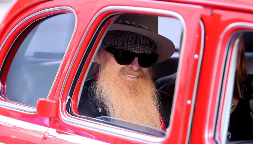 Billy Gibbons, jeden z członków ZZ Top, w stylowym kapeluszu i z charakterystyczną brodą, siedzi w czerwonym samochodzie.