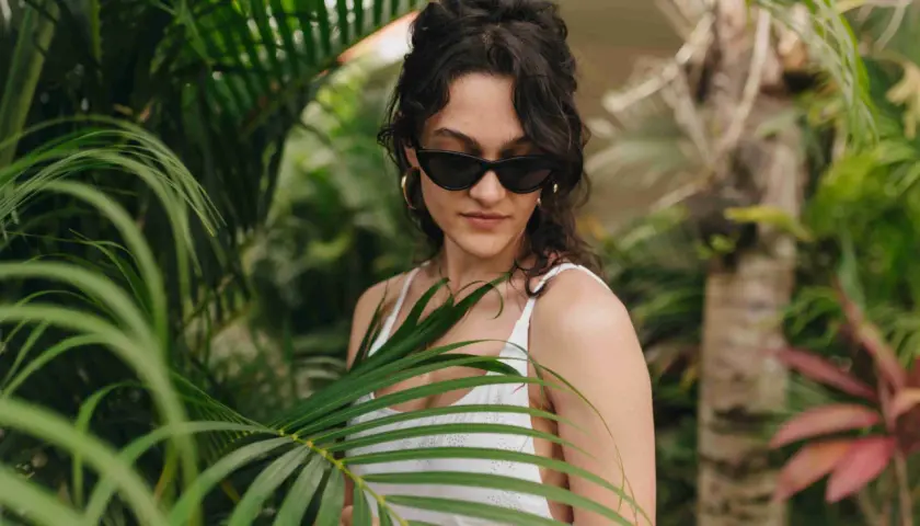 Joven con gafas de sol y traje de baño blanco entre hojas verdes, un ejemplo de moda ecológica.