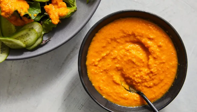A vibrant bowl of carrot ginger dressing sits next to a salad with cucumber slices.