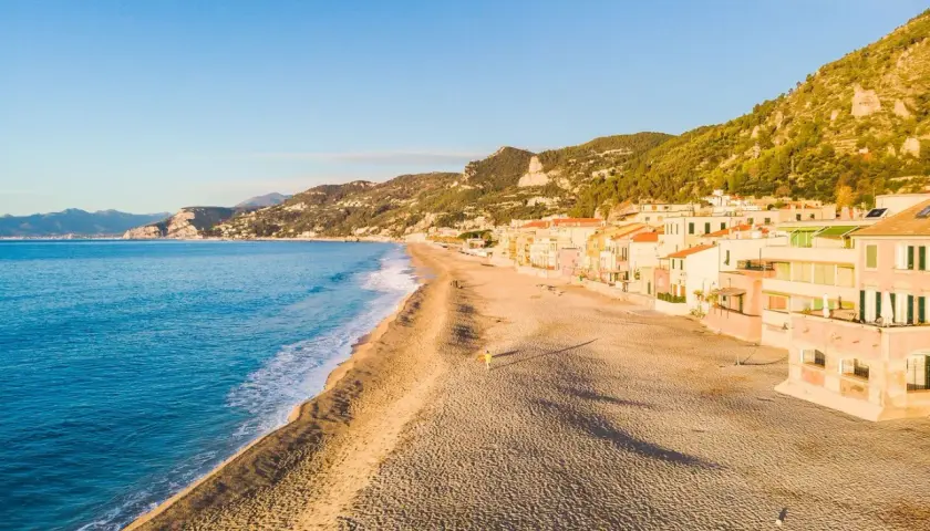 Plage de sable fin bordée de maisons colorées sur la côte italienne. La mer bleue et les collines verdoyantes complètent ce paysage paisible.