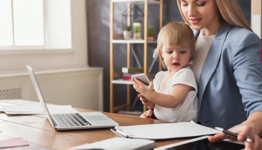 Kobieta z dzieckiem przy biurku z laptopem i tabletami. Czy na urlopie macierzyńskim można pracować na umowę zlecenie?