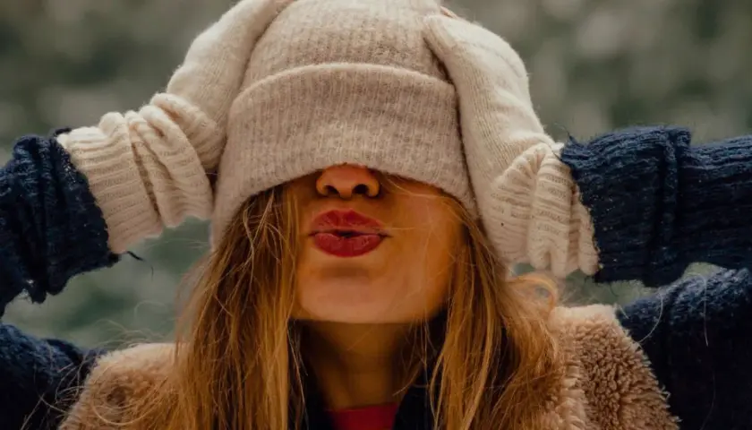 Une jeune femme s'amuse à s'habiller chaudement pour affronter le froid, cachant son visage sous un bonnet et des moufles.