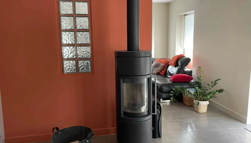 Un poêle moderne noir se dresse devant une peinture murale ocre, à côté d'une fenêtre en verre dépoli. Un canapé en cuir noir et des coussins colorés sont visibles au loin.