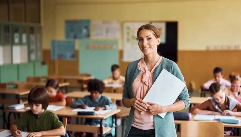 Lächelnde Lehrerin mit Unterlagen in einem Klassenzimmer voller konzentrierter Kinder. Sie fragt sich: "Soll ich Lehrer werden?