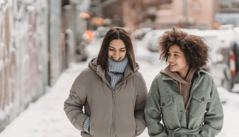Deux amies profitent d'une promenade hivernale, bien emmitouflées dans leur vêtement contre le froid. Le top 7 des tenues chaudes pour affronter la neige.