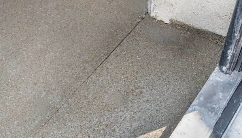 Szara, ziarnista powierzchnia, prawdopodobnie beton, z widocznym pęknięciem. Obok biała ściana i fragment czarnej poręczy. Mastyka użyta do uszczelnienia.