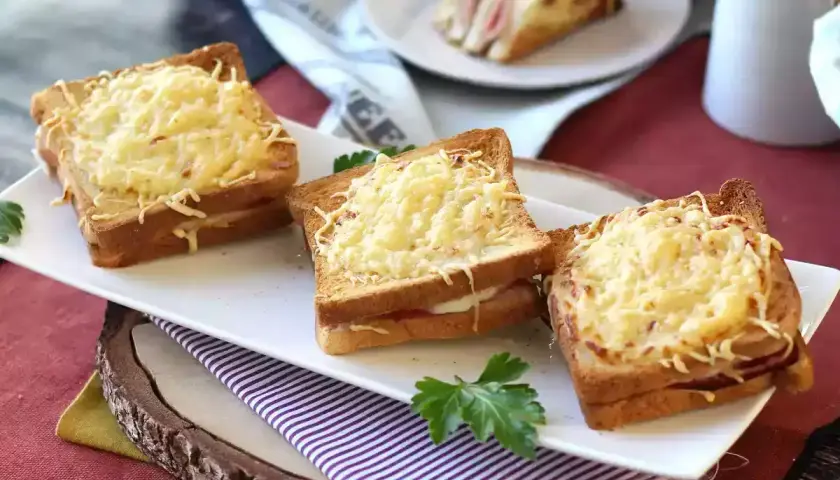 Tre gyllene croque monsieur, gratinerade med ost, ligger uppradade på en vit bricka. En klassisk fransk delikatess.