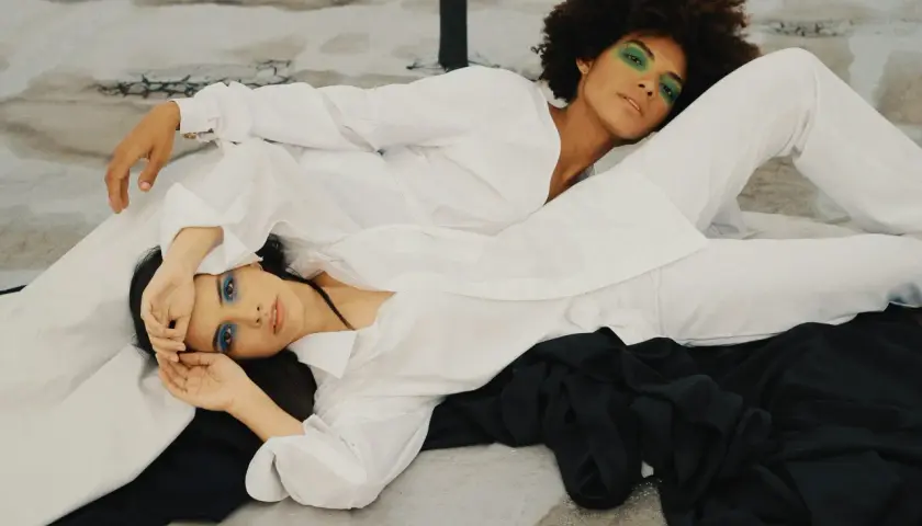 Two women in white shirts and pants, one with green eyeshadow, the other with blue. The fabric looks smooth, suggesting lyocell does not wrinkle easily.