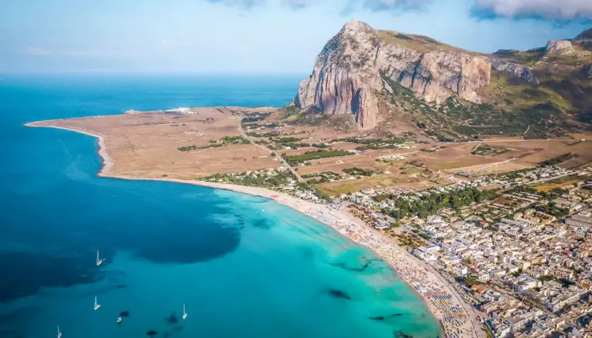 Aerial view of a crowded beach and town, showcasing one of the best beach towns in Sicily with turquoise waters and a dramatic mountain backdrop.