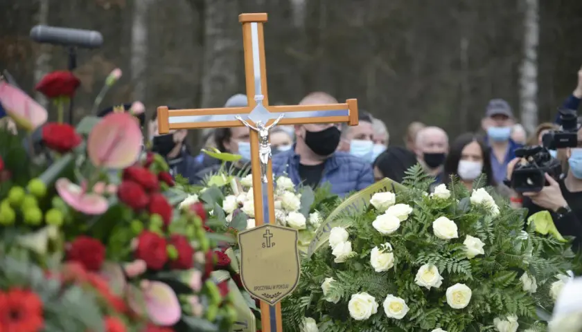 Drewniany krzyż z figurą Chrystusa na pogrzebie. Wieniec białych róż i czerwonych kwiatów. Ludzie w maseczkach. Co zrobić z krzyżem po pogrzebie?