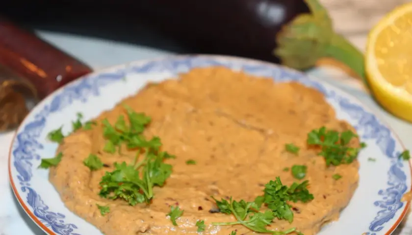 En krämig baba ganoush, garnerad med persilja, med aubergine och citron i bakgrunden.