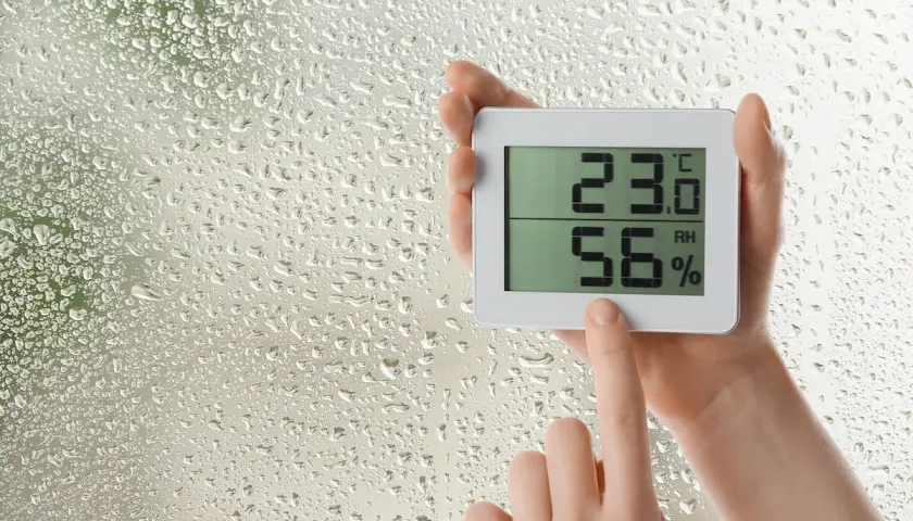 Manos sostienen un higrómetro digital que muestra 23.0°C y 56% de humedad, mientras gotas de lluvia cubren el cristal.