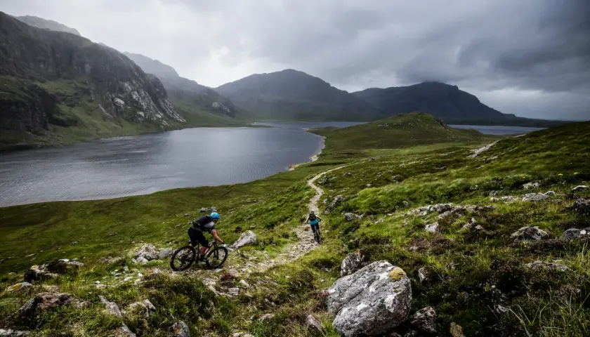 Two cyclists navigate a rocky trail with a vast lake and dramatic mountains behind them, showcasing the rugged beauty of Scotland bike tours.