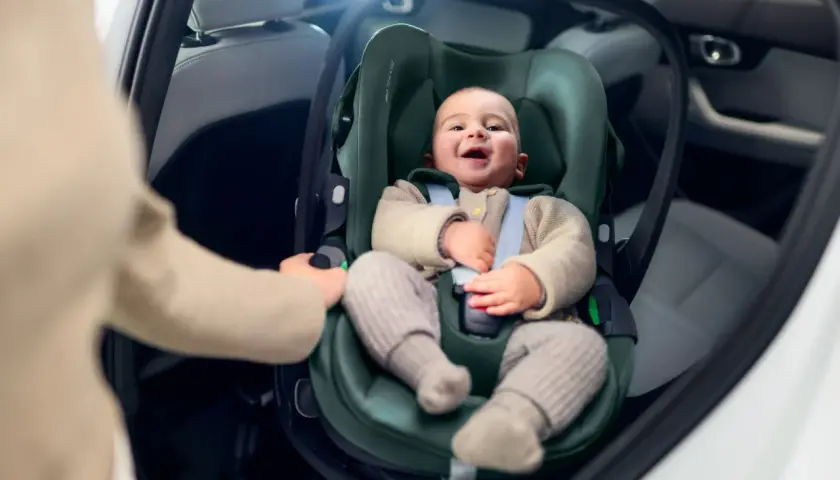 Uśmiechnięte dziecko w foteliku samochodowym. Ręka rodzica pokazuje, jak wypiąć fotelik z bazy isofix.