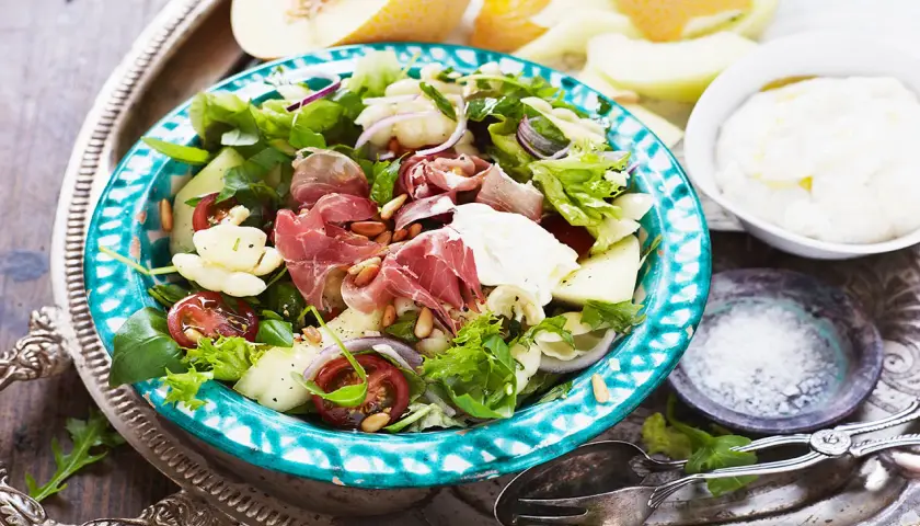 En härlig ost och skinksallad med melon, tomater och pinjenötter. Serveras på en vacker turkosblå skål.
