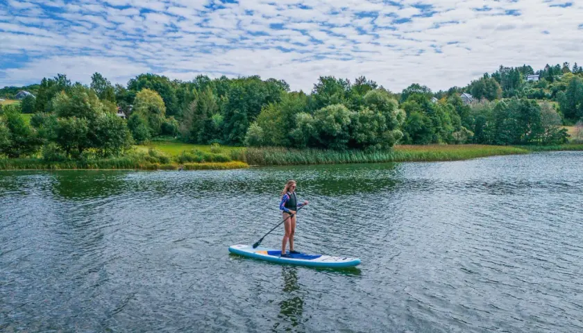 Kobieta na desce SUP na spokojnym jeziorze, w tle zielone drzewa i błękitne niebo.