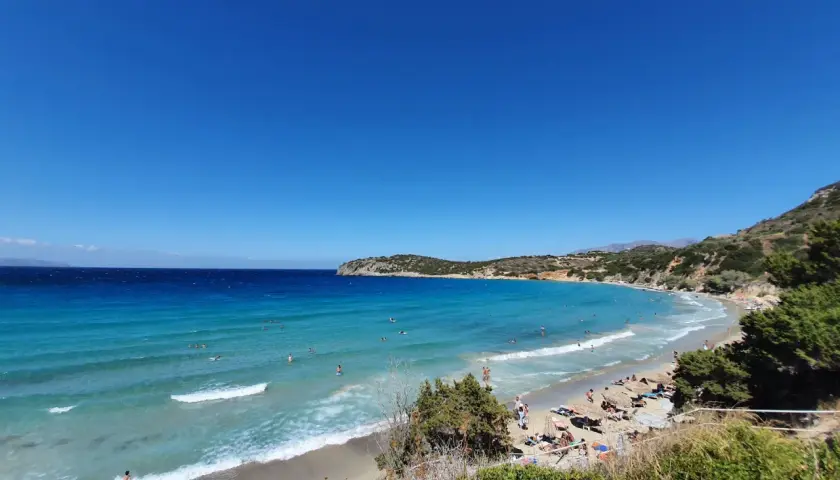 Słoneczny dzień na plaży Voulisma. Turkusowa woda, piaszczysty brzeg i zielone wzgórza tworzą idylliczny krajobraz.