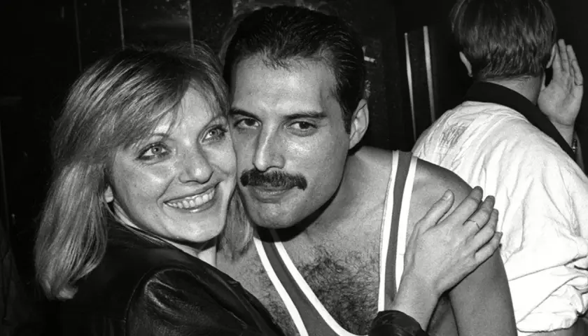Freddie Mercury z uśmiechem, odsłaniającym jego charakterystyczne zęby, przytula się do kobiety.