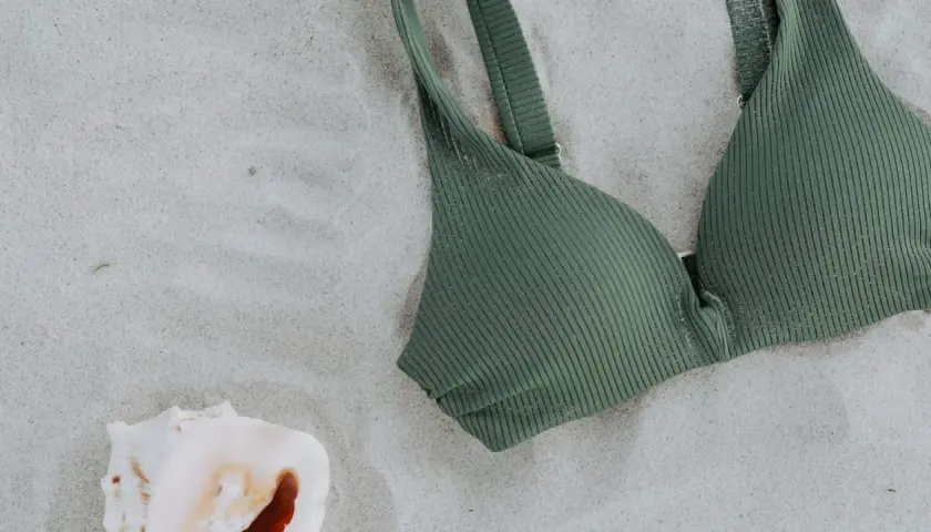 A ribbed green swim fabric bikini top rests on the sand next to a seashell.