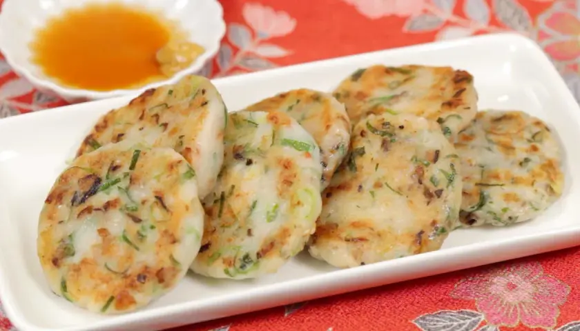 Golden-brown daikon mochi patties, studded with green onions, are served on a white platter with a small dish of dipping sauce.