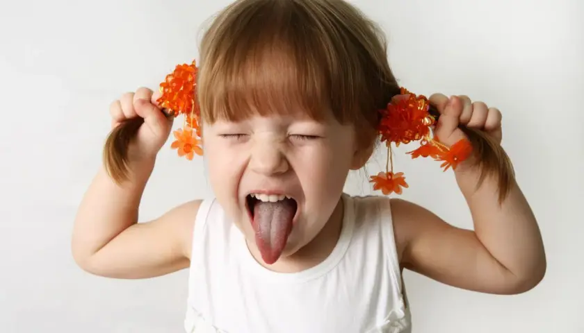 Bambina iperattiva fa una smorfia con la lingua fuori, tirando le trecce con fiori arancioni. Cosa fare?