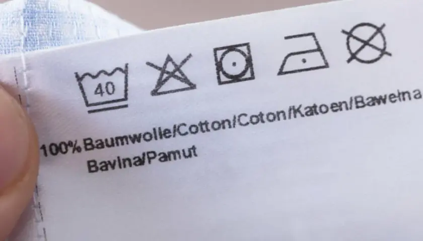 Pflegeetikett mit Waschsymbolen und dem Hinweis "100% Baumwolle". Die Symbole zeigen, dass das Kleidungsstück bei 40°C waschbar ist, nicht bleichen, bei niedriger Temperatur im Trockner trocknen und bei mittlerer Hitze bügeln.