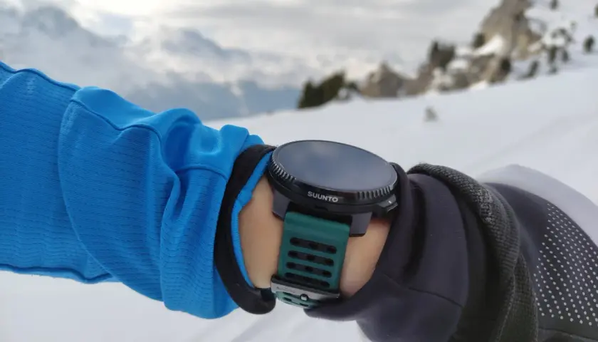 Suunto Vertical Test: Arm in blauer Jacke mit grüner Suunto-Uhr im Schnee, Bergkulisse.