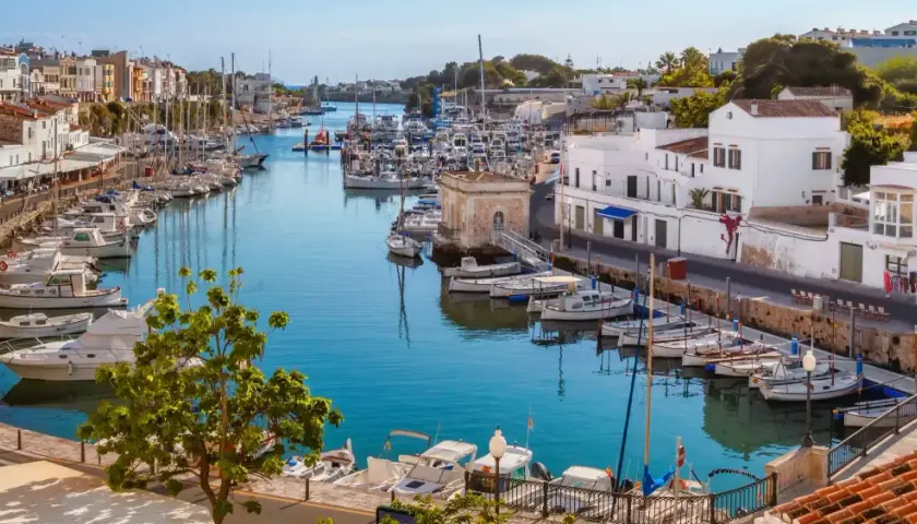 Hafen von Ciutadella, Menorca. Boote im türkisfarbenen Wasser, weiße Häuser und bunte Fassaden. Ein malerischer Blick auf die Menorca Highlights.