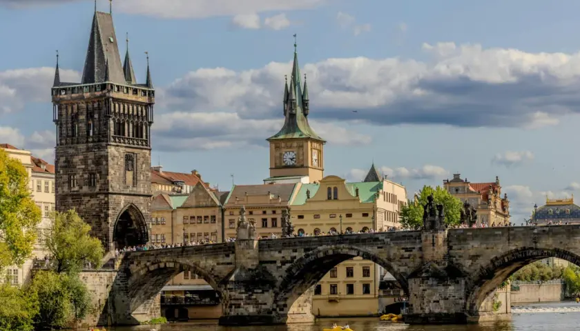 Most piękne stolice Europy, Most Karola w Pradze, z wieżami i zegarem, na tle błękitnego nieba z chmurami.