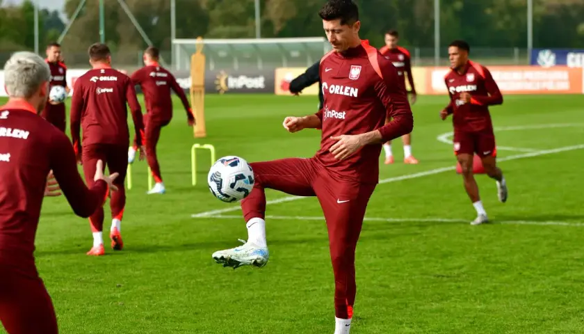 Piłkarze w bordowych strojach ćwiczą na boisku. Robert Lewandowski bawi się piłką, pokazując swoje umiejętności w najpopularniejszym sporcie na świecie.