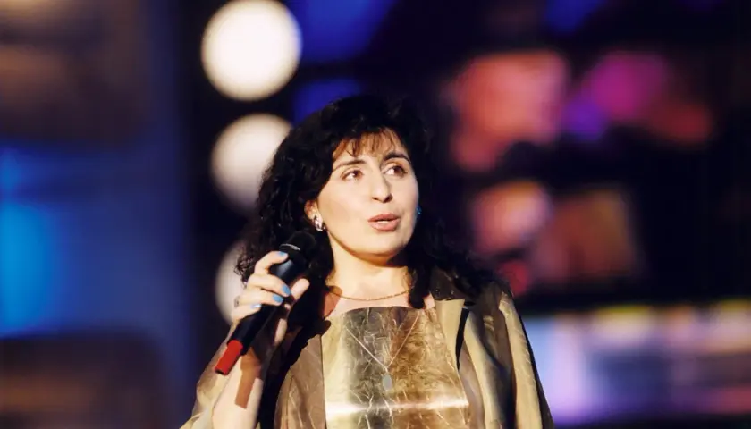 Eleni śpiewa na scenie, trzymając mikrofon.