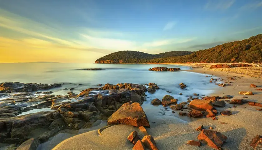 Coucher de soleil sur une plage rocheuse en Maremma, Italie. L'eau calme caresse le sable doré et les rochers ocre sous un ciel aux teintes chaudes.