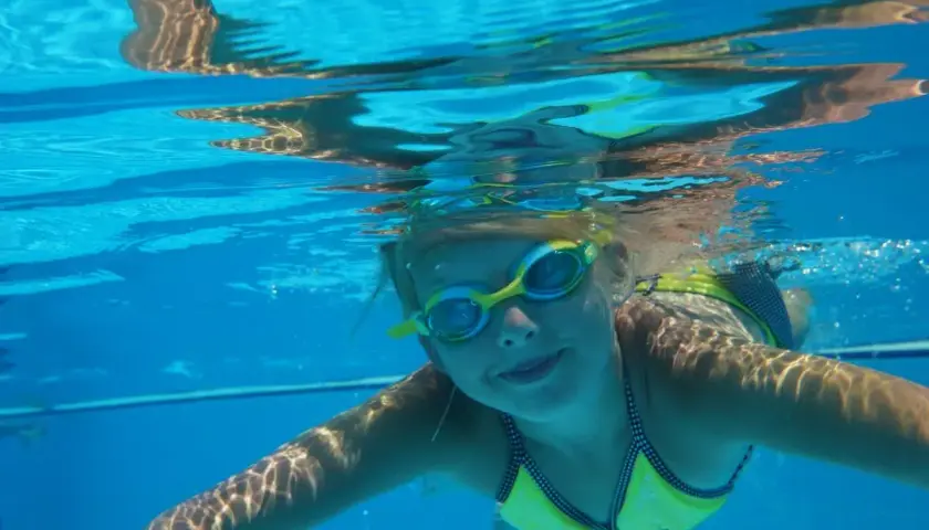 Enfant souriant sous l'eau, lunettes de piscine, maillot jaune. Attention à l'otite de piscine après ces moments joyeux.