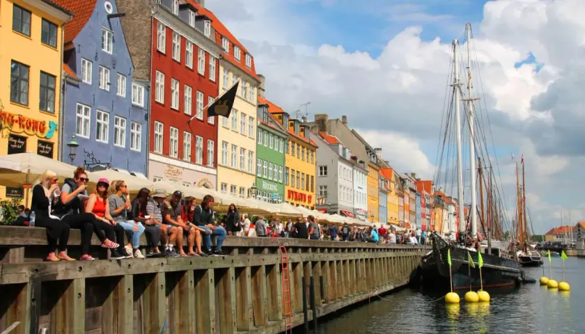 Bunte Häuser am Nyhavn, Kopenhagen an einem Tag. Menschen sitzen am Kai, Boote liegen im Wasser.