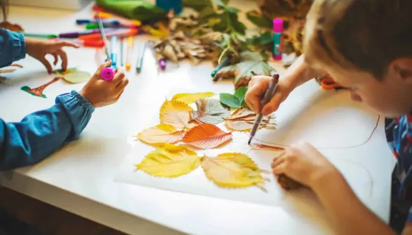 Niños de otoño creando arte con hojas secas y rotuladores.