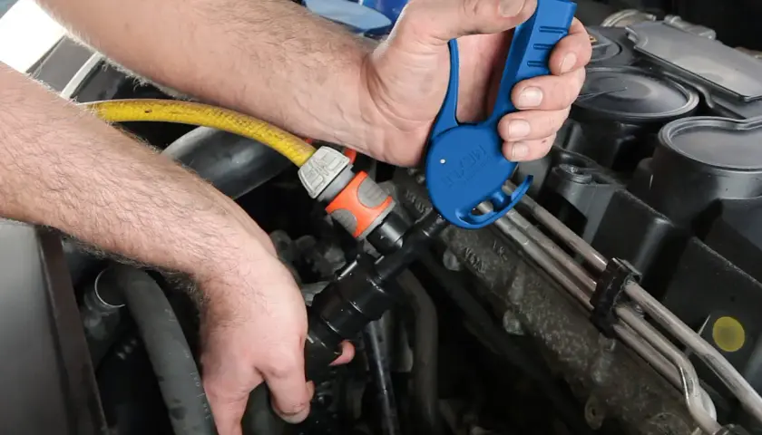Ręce mechanika podłączają niebieskie narzędzie do węża, przygotowując środek do płukania układu chłodzenia.
