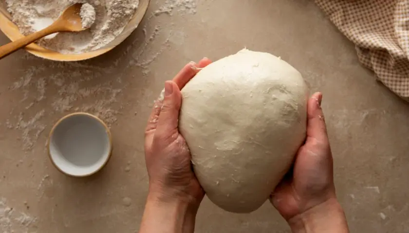 Mani che impastano un panetto soffice, ingrediente chiave per imparare come fare il lievito madre. Farina e ciotolina a lato.