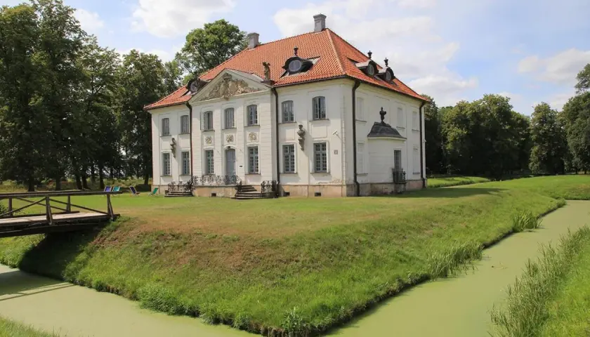 Pałac w Choroszczy otoczony fosą i zielenią, z drewnianym mostkiem prowadzącym do rezydencji.