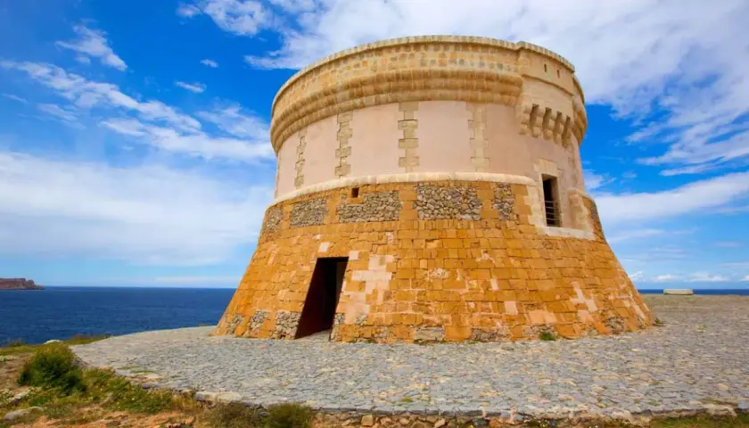 Torre de defensa costera, un lugar histórico que ver en Fornells. El mar azul y el cielo nublado completan la vista.
