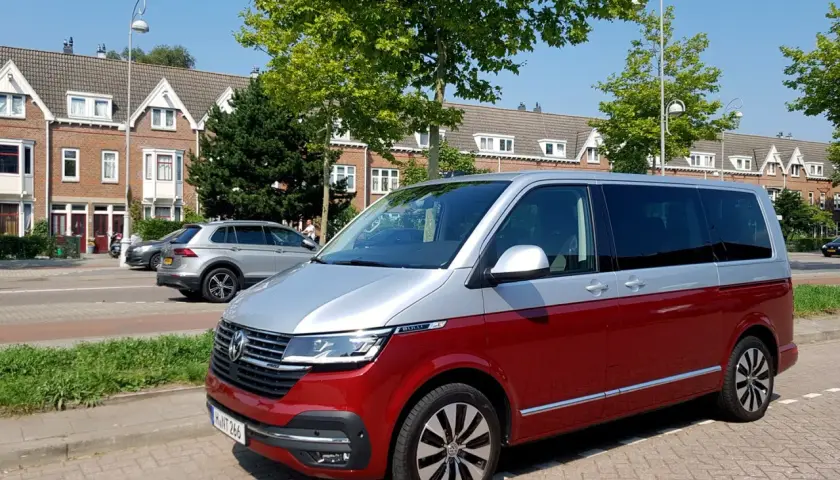 Dwukolorowy volkswagen van, czerwono-srebrny, zaparkowany na tle domów i drzew.
