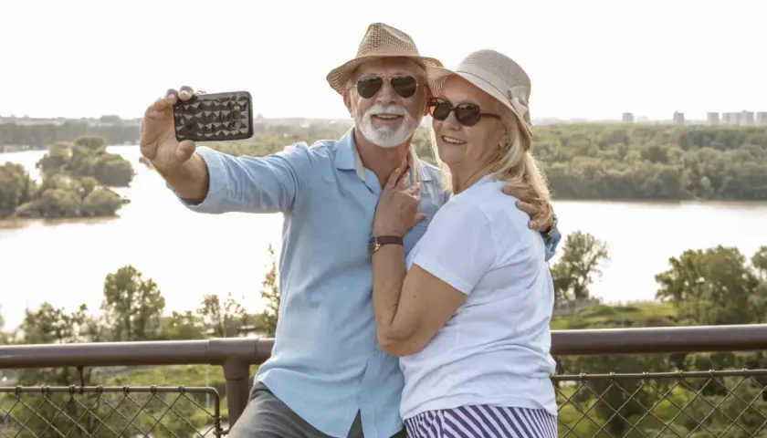 Uśmiechnięta para seniorów robi selfie na tle rzeki. Idealne wycieczki dla seniorów samolotem to świetny sposób na podróżowanie.