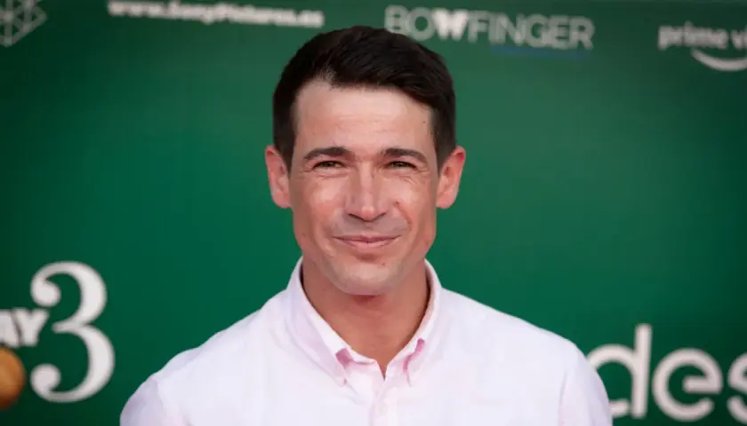 Juan José Ballesta, cuya edad se refleja en su mirada, sonríe con camisa rosa ante un fondo verde con logos.