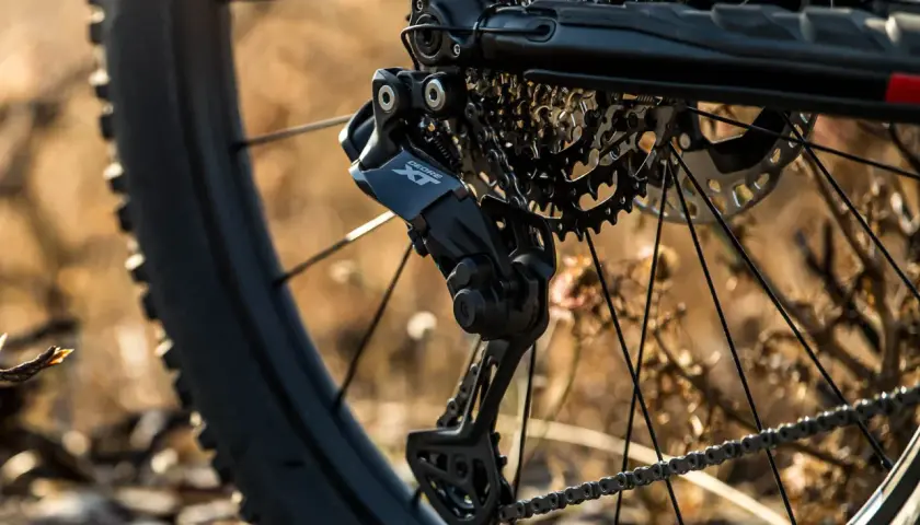 Close-up of a bicycle's rear derailleur, showcasing the Shimano Deore XT component, hinting at advanced Shimano wireless shifting technology.