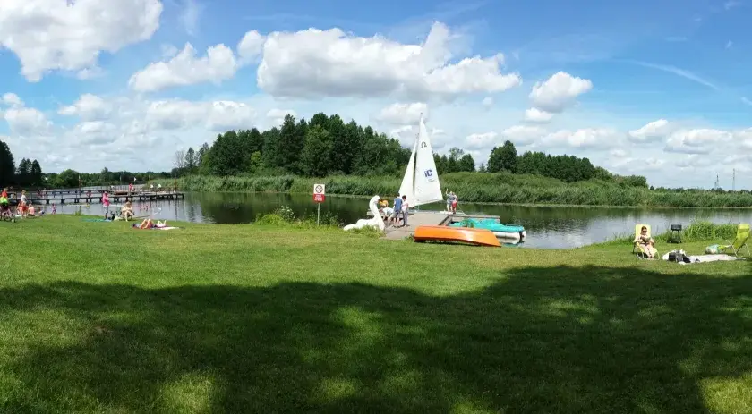 Słoneczny dzień nad jeziorem Kozienickim. Ludzie odpoczywają na trawie, opalają się, a na wodzie pływa żaglówka.